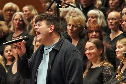 Ein Mann mit dunklen Haaren steht vor einem Chor und singt mit Inbrunst in ein Mikrofon. - Copyright: Dirk Tostmann